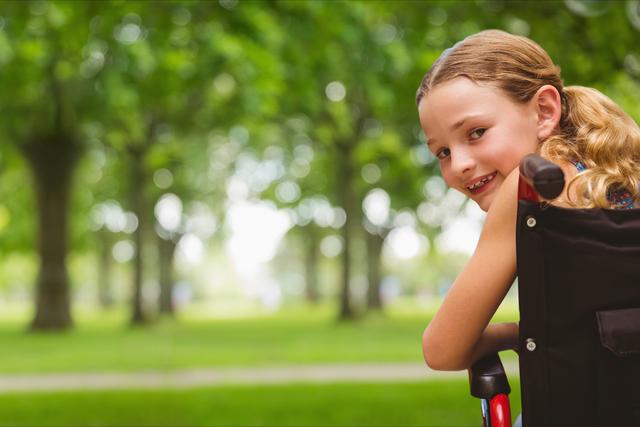 Young girl sitting in a wheelchair at a park, smiling while looking back at the camera. Surrounded by lush green trees and open grassy areas, she embodies happiness and inclusion. Perfect for concepts related to accessible environments, childhood joy, inclusivity, and support for those with mobility challenges.