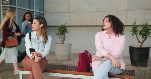 Two diverse women sitting on bench in urban courtyard holding phone and looking away
