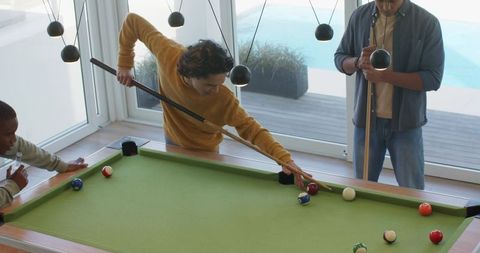 Multicultural friends playing pool on green-felt table in contemporary minimalist rec room