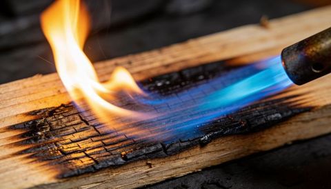 Blowtorch charring wood plank with blue flame creating cracked char texture