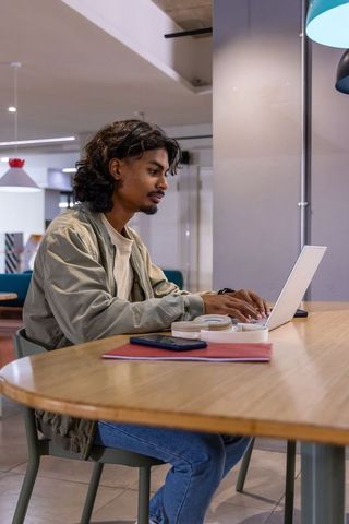 Indian man working on laptop in modern coworking lounge with notebook and phone