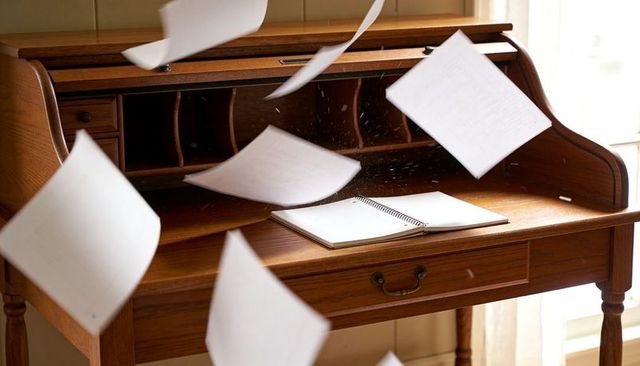 Vintage roll-top desk with papers flying and open spiral notebook in warm sunlight