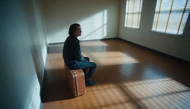 Solitary traveler sitting on vintage suitcase in empty sunlit room with window shadows
