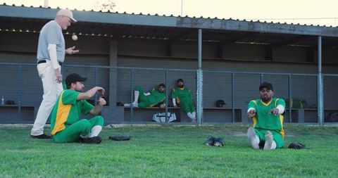 Coach tossing ball while baseball players stretching near dugout in green uniforms