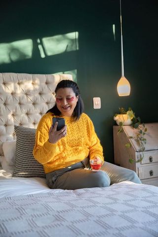 Smiling woman sitting on bed wearing yellow sweater holding phone and drink, cozy bedroom