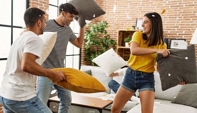 Young adults having pillow fight in sunlit loft living room with mustard and gray cushions