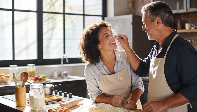 Middle-aged couple baking in rustic kitchen, smiling, man sprinkling flour on partner