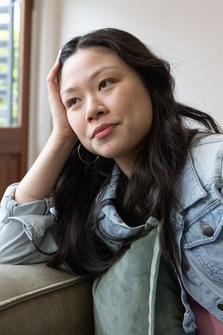 Asian woman gazing thoughtfully on sofa in denim jacket, casual lifestyle portrait