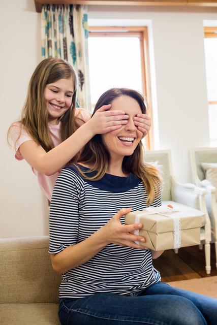 Daughter covering her mother's eyes while giving a surprise gift in a cozy living room. Perfect for themes related to family bonding, celebrations, parenting, and joyful moments at home.