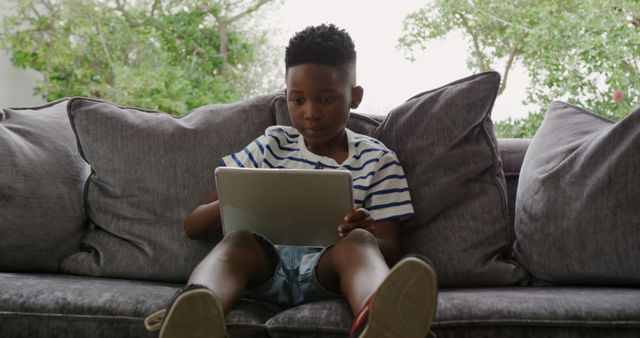 Image shows young boy sitting on a gray sofa in a cozy living room, using a digital tablet. The casual and relaxed atmosphere suggests a home or comfortable indoor environment. The boy is wearing a striped shirt and has afro hair, adding to the casual look. Greenery seen through the background window brings a sense of freshness to the space. Perfect for websites and articles about childhood, technology use, indoor activities, and casual lifestyle content.