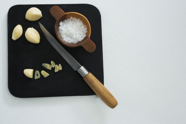 This image shows chopped garlic cloves and a bowl of sea salt on a black chopping board, accompanied by a knife with a wooden handle. Ideal for use in culinary blogs, recipe websites, cooking tutorials, and food-related advertisements. It highlights the preparation and seasoning process in cooking.