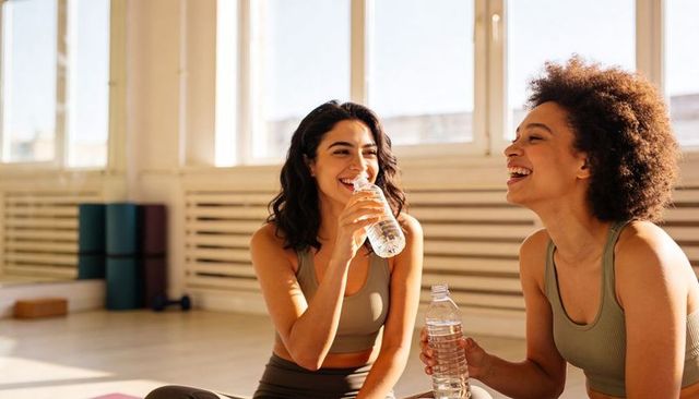 Two women laughing and drinking water after yoga class in sunlit studio post-workout refresh