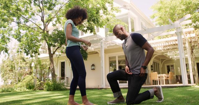 Man kneeling with engagement ring proposing to woman outside a suburban house in a lush green garden. Ideal for advertisements celebrating love, relationship counseling, engagement party invitations, or romantic occasions. Perfect for depicting romantic moments in outdoor settings.