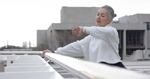 Senior woman checking watch while leaning on railing on brutalist rooftop terrace