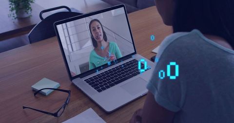 Watching remote video consultation on laptop at wooden desk with glasses and notepad