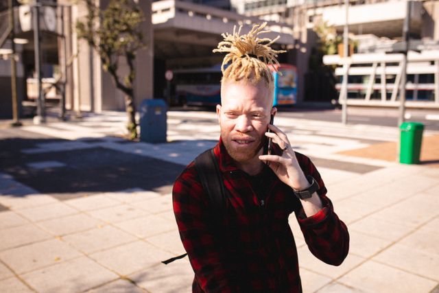 Albino young man with dreadlocks talking on smartphone outdoors in urban setting. He is wearing a red plaid shirt and carrying a backpack. This image can be used for topics related to albinism, skin conditions, modern communication, urban lifestyle, and youth culture.