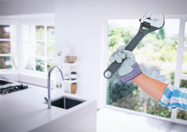Hand wearing glove holding adjustable wrench in modern kitchen. Ideal for illustrating home improvement, DIY projects, plumbing repairs, and maintenance tasks. Bright and clean kitchen setting emphasizes the importance of keeping home spaces functional and well-maintained.