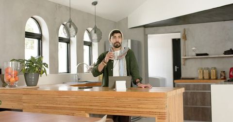 Young indian man standing at wooden kitchen island sipping warm coffee and viewing tablet