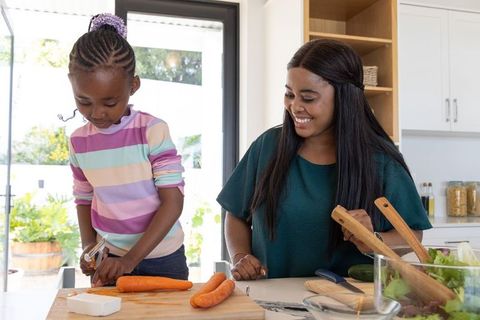 African american mom and daughter peeling carrots in bright kitchen, cooking bonding