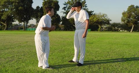 Two Cricket Teammates Chatting and Adjusting Cap on Sunlit Grass Field During Practice