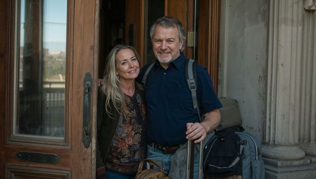 Senior couple smiling and leaving historic doorway with luggage for urban getaway