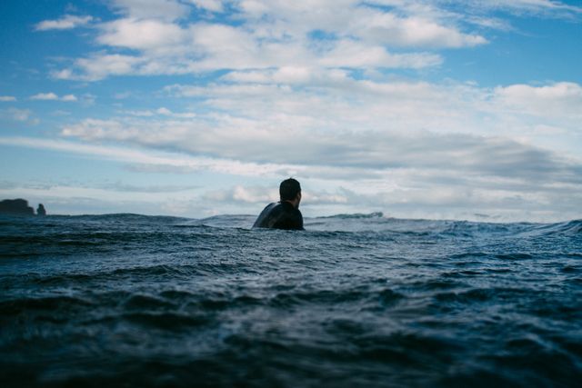 Surfer waiting for the perfect wave in tranquil ocean, a person partially submerged in the water with a cloudy sky. Great for use in projects related to surfing, water sports, adventure, and outdoor activities.