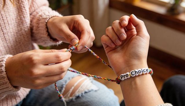Tying multicolored friendship bracelet with pro-choice beads, hands braiding closeup, cozy