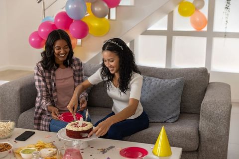 Multiracial friends cutting red velvet cake on sofa during colorful birthday celebration