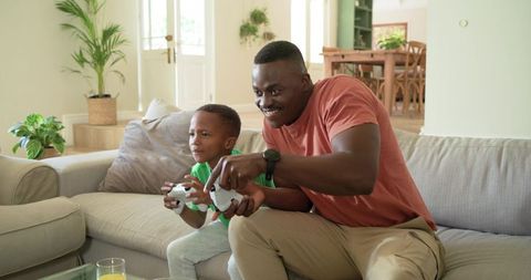 African American Father and Son Playing Video Games on Couch, Smiling, Home Bonding