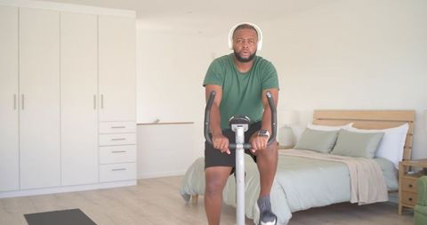 Man exercising on stationary bike in minimalist bedroom wearing headphones and smartwatch