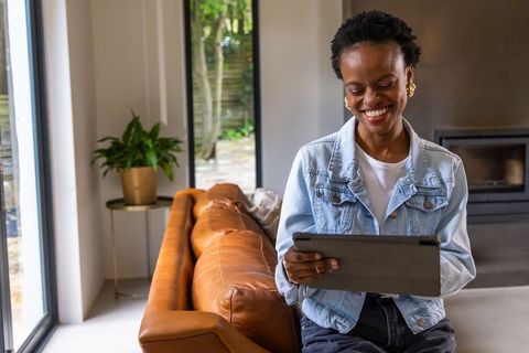Smiling african american woman using tablet on leather sofa in modern living room