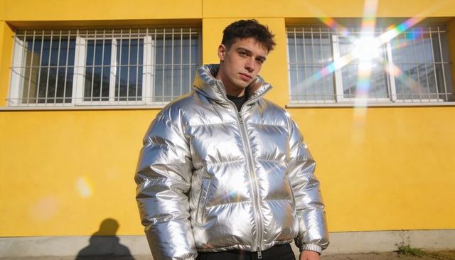 Young man posing in silver metallic puffer jacket against yellow building with sun flare