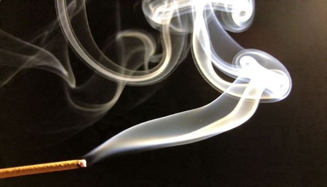 Incense smoke spirals releasing ethereal ribbons over dark background with glowing ember