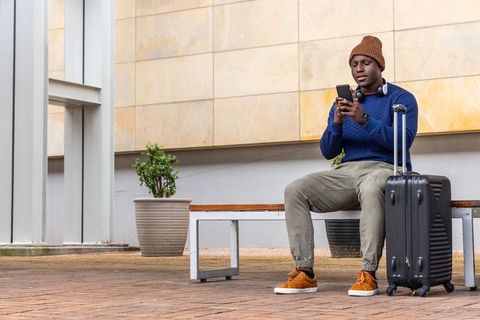 Young traveler sitting on bench checking phone with suitcase and headphones in urban plaza