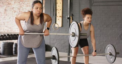 Two women performing bent-over barbell rows wearing grey tanks and smartwatches in industrial gym
