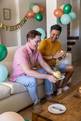 Diverse male friends celebrating birthday at home with cake, balloons and smiles