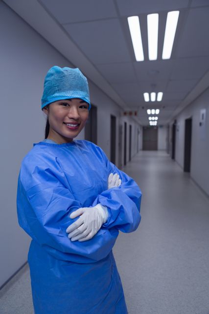 Female surgeon wearing blue scrubs and gloves, standing with arms crossed in a hospital corridor. Image conveys confidence, professionalism, and the medical environment. Ideal for healthcare articles, hospital promotional materials, and medical staffing websites.