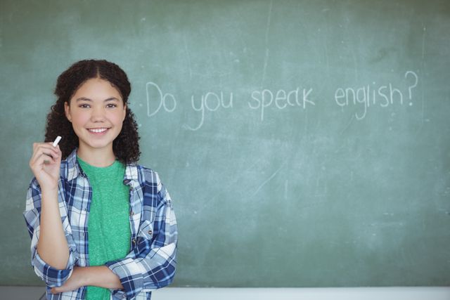 Smiling schoolgirl with curly hair stands in front of a chalkboard holding chalk, pretending to be a teacher. The chalkboard has the question 'Do you speak English?' written on it. This image is perfect for educational materials, back-to-school promotions, and articles about childhood learning and development.