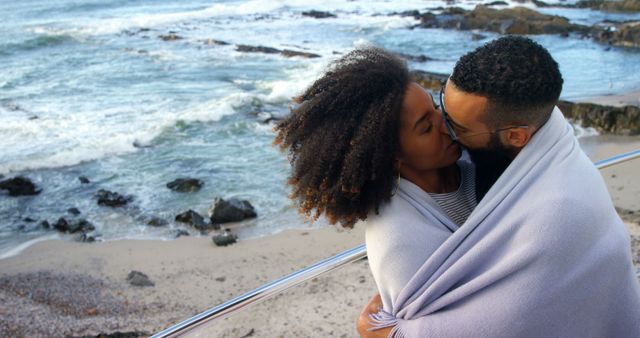 Biracial couple in love kissing wrapped in blanket by seaside with copy space. Love, relationship, vacation and lifestyle concept, unaltered.
