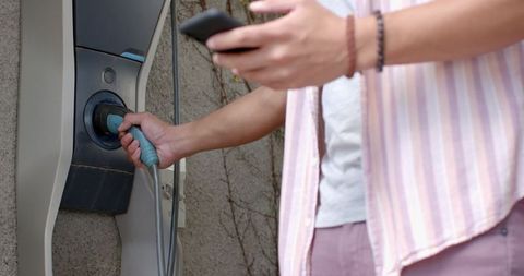 Person charging electric vehicle and using smartphone