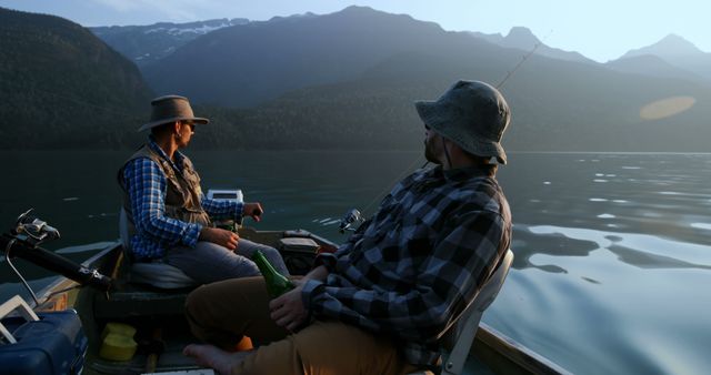 Two friends enjoy a peaceful fishing trip on a mountain lake at sunset. The surrounding mountains and still water create a serene atmosphere. Ideal for content related to outdoor activities, travel adventures, relaxation, bonding experiences, or promotional material for fishing equipment and apparel company.