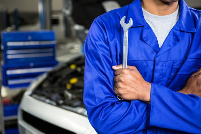 Mid section of mechanic with arms crossed holding spanner at repair garage
