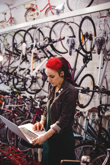 A female bicycle mechanic with red hair using a laptop in a workshop with bicycles hanging on the wall behind her. This image can be used to depict a modern bike repair shop, technology in maintenance work, female professionals in technical fields, or a small business environment.
