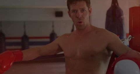 Shirtless boxer leaning on ropes wearing red gloves in dim gym corner