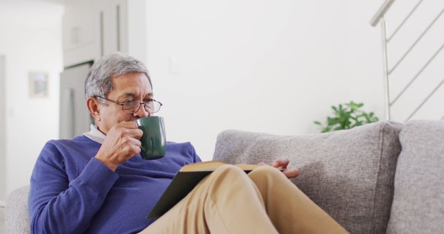 Senior man enjoying a relaxing moment in living room, sipping a cup of coffee while reading a book on a couch. Perfect for depicting themes of leisure, retirement, relaxing at home, mature lifestyles, and cozy indoor settings.