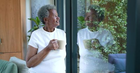 Senior african woman holding mug and looking out window, reflection of greenery, peaceful moment