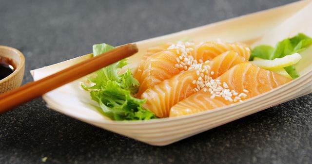 Fresh salmon sashimi in wooden tray with sesame seeds, green lettuce, and soy sauce in background. Ideal for illustrating Japanese cuisine, healthy eating, gourmet dining, and seafood recipes.