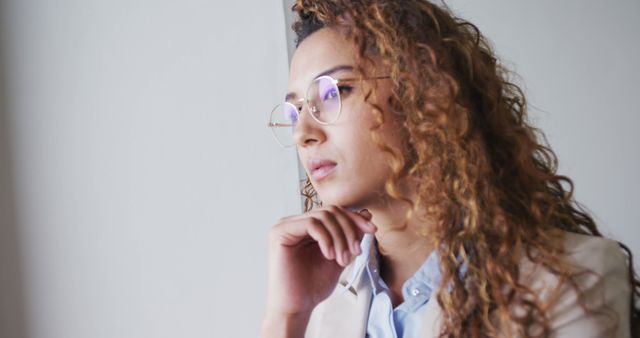 Mixed-race businesswoman wearing eyeglasses and business attire looking thoughtful with hand on chin. Useful for advertisements, professional career promotions, articles on business women, diversity in workplace, mental well-being at work, and intelligent, modern professionals.