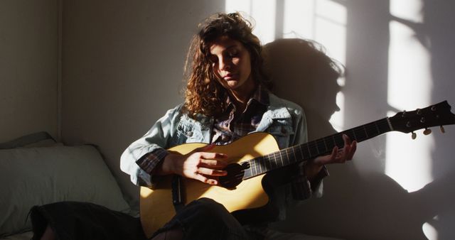 Relaxed biracial woman sitting and playing acoustic guitar in sunny cottage bedroom. simple living in off the grid rural home.