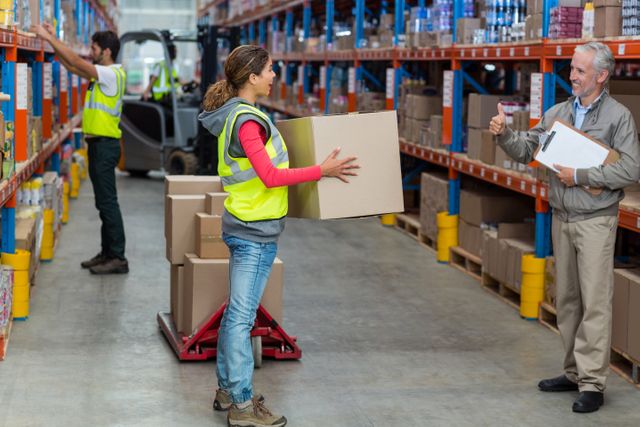 Warehouse manager showing thumbs up to female worker while carrying cardboard boxes in warehouse